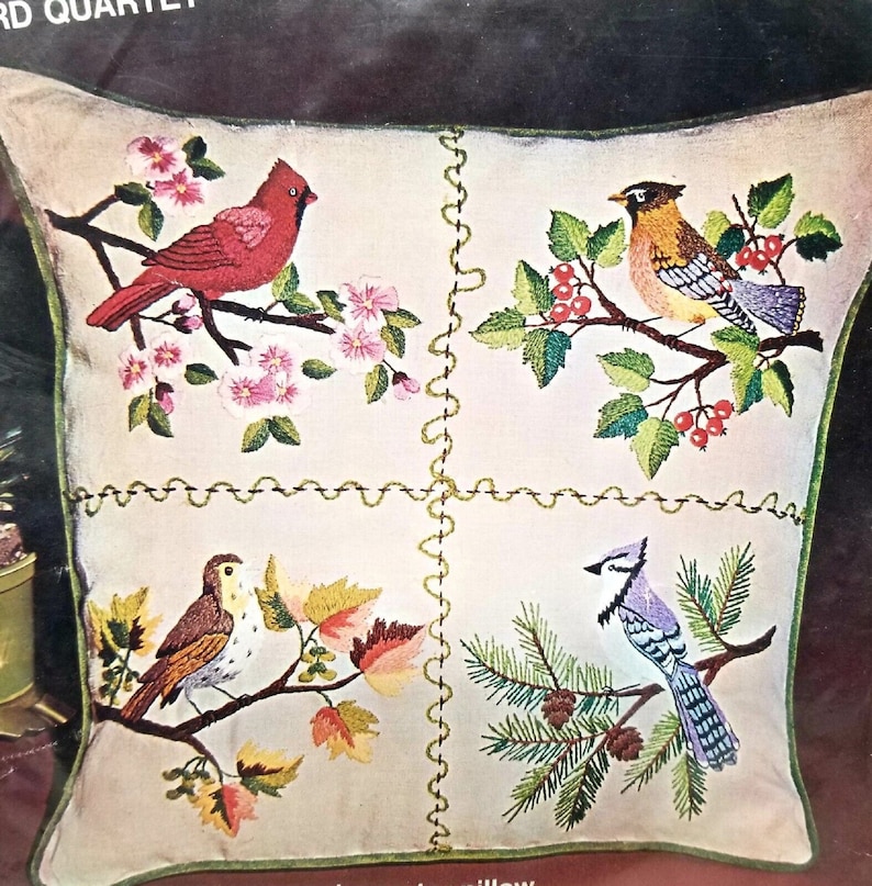 May include: A white pillow with four embroidered squares featuring different birds and foliage. The top left square shows a red cardinal on a branch with pink blossoms. The top right square shows a robin on a branch with green leaves and red berries. The bottom left square shows a brown bird on a branch with fall leaves. The bottom right square shows a blue jay on a branch with pine needles and pine cones.