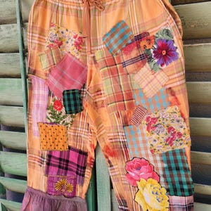 May include: Orange plaid pants with colorful patchwork details. The pants feature a drawstring waist, ruffled hems, and various floral and plaid patches in shades of pink, green, and yellow. The pants are hanging on a green shutter.