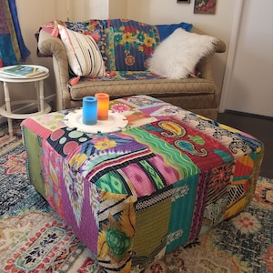May include: A colorful patchwork ottoman with a variety of fabric patterns and colors, including paisley and floral designs. The ottoman is square and sits on a patterned rug. Two candles sit on a white decorative plate on top of the ottoman.