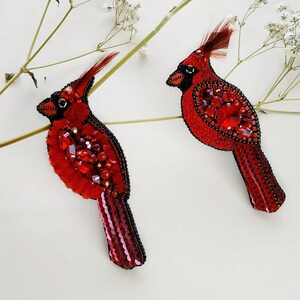 Puede incluir: Dos broches de pájaro cardenal rojo. Los pájaros están adornados con cuentas rojas, lentejuelas y plumas. Los broches tienen contornos de cuentas negras y están sobre un fondo blanco.