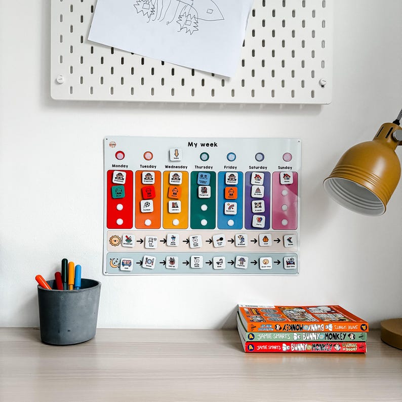 May include: A white magnetic board with a colorful daily schedule for kids. The board has a section for each day of the week, with spaces for magnetic icons. The board is labeled "My week".