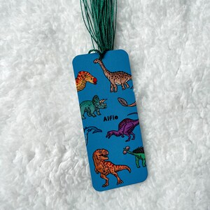 May include: Blue bookmark featuring a range of dinosaur illustrations, including a T-Rex, a Triceratops, and a Stegosaurus. The bookmark also has the name "Alfie" printed on it.