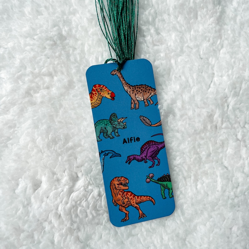 May include: Blue bookmark featuring a range of dinosaur illustrations, including a T-Rex, a Triceratops, and a Stegosaurus. The bookmark also has the name "Alfie" printed on it.