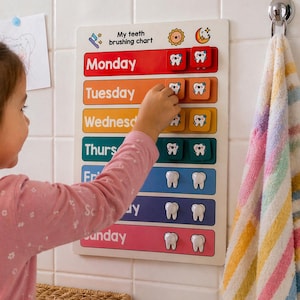 Op de afbeelding: Een tandenpoetskaart voor kinderen met de woorden "My teeth brushing chart" bovenaan. De kaart heeft kleurrijke secties voor elke dag van de week, met magnetische tandvormige pictogrammen. Een kind plaatst een tandpictogram op de kaart.