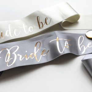 Silver bachelorette sash with metallic gold writing. It is open ended and comes with a heart shaped acrylic pin to secure the ends. The ribbon is 66 mm wide and 180 cm long. Colours of the ribbon and the text are customizable.