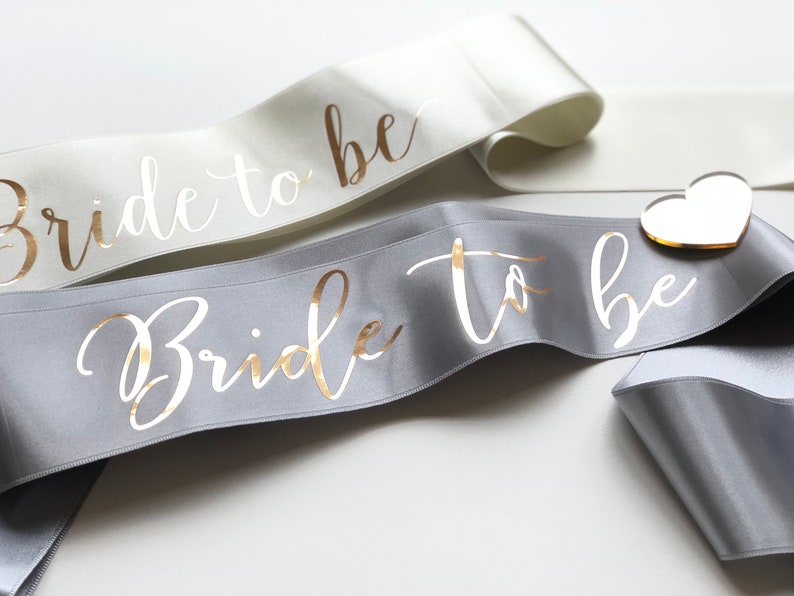 Silver bachelorette sash with metallic gold writing. It is open ended and comes with a heart shaped acrylic pin to secure the ends. The ribbon is 66 mm wide and 180 cm long. Colours of the ribbon and the text are customizable.