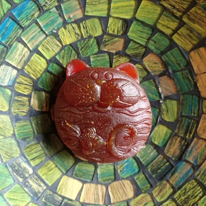 May include: A round, reddish-brown soap with a cat face design, set in a mosaic bowl with green, blue, and yellow tiles. The soap has a detailed carving of a fish and other patterns.