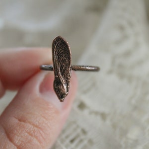 May include: A copper ring with a detailed maple seed design. The ring is worn on a finger.