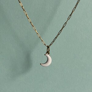 May include: A gold-toned chain necklace featuring a small, white crescent moon pendant. The pendant is attached to the chain by a simple gold-toned jump ring. The chain consists of elongated, rectangular links connected in a continuous pattern.