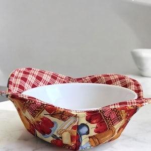 May include: A white ceramic bowl nested in a colorful fabric bowl cozy. The cozy features a pattern of red and yellow kitchen utensils and food items on a beige background, with a red and white plaid pattern on the rim.