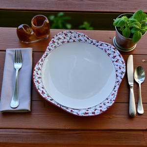 May include: A white plate with a cherry-patterned rim sits on a wooden table, next to a fork, knife, and spoon. A small pot of basil and a glass condiment holder are also on the table. The plate is surrounded by a red border.