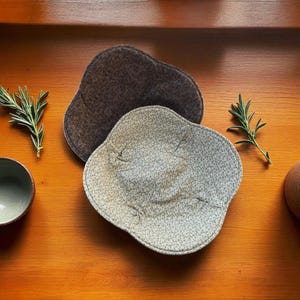 May include: Two handmade, irregularly shaped fabric bowls. One is a light gray with a small floral pattern, and the other is a solid dark brown. The bowls are on a wooden surface, with sprigs of rosemary and a small ceramic bowl.