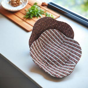 May include: Two heart-shaped fabric placemats. One features a brown floral pattern, and the other has a striped design with white, pink, and brown stripes. The placemats are placed on a light-colored surface.