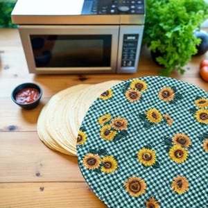 May include: A round, quilted placemat with a green and white checkered pattern and yellow sunflowers. A stack of tortillas, a small bowl of red sauce, and a microwave oven are also visible on a wooden surface.