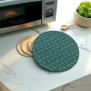 May include: A green, round, floral-patterned hot pad next to a stack of tortillas. A microwave oven and a small wooden spoon are also visible. The hot pad has a repeating pattern of small pink flowers and white accents.