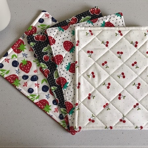 May include: Set of four quilted fabric pot holders with fruit patterns. Designs include strawberries, cherries, and blueberries on white and black backgrounds. The pot holders are square and diamond-stitched.