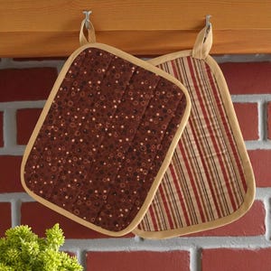 May include: Two quilted pot holders hanging on hooks. One is brown with a pattern of white and brown circles. The other has red, brown, and white stripes. Both have tan borders and hanging loops. The background is a brick wall.
