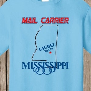 May include: Light blue t-shirt with a black outline of the state of Mississippi and the text "MAIL CARRIER" in red and black. The text "LAUREL 39440" is in red and black, and the text "MISSISSIPPI" is in blue and black. The t-shirt has a white tag with the text "PORT & COMPANY" on it.