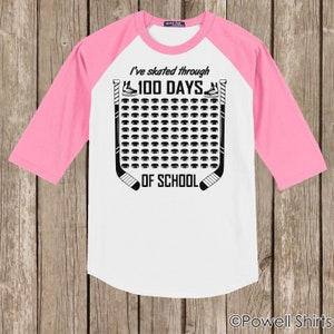 K&ouml;nnte beinhalten: Wei&szlig;es und rosafarbenes Baseball-T-Shirt mit schwarzem Text, der "Ich bin 100 Tage lang durch die Schule gelaufen" lautet. Der Text ist von einem Hockey-Schl&auml;ger- und Puck-Design umgeben.