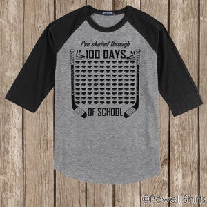 K&ouml;nnte beinhalten: Ein graues und schwarzes Baseball-Raglan-T-Shirt mit dem Text "I've skated through 100 days of school" und einer Grafik mit einem Hockeyschl&auml;ger und einem Puck.