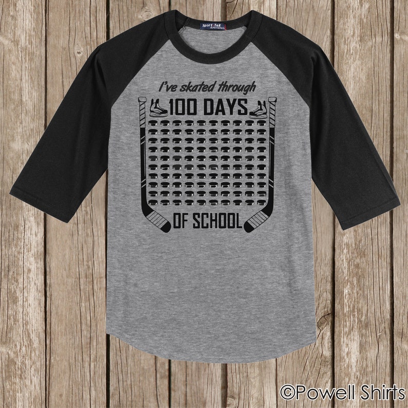 K&ouml;nnte beinhalten: Ein graues und schwarzes Baseball-Raglan-T-Shirt mit dem Text "I've skated through 100 days of school" und einer Grafik mit einem Hockeyschl&auml;ger und einem Puck.