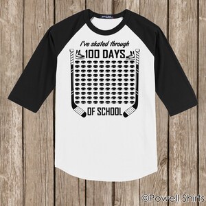 K&ouml;nnte beinhalten: Ein wei&szlig;-schwarzes Baseball-T-Shirt mit einem Hockey-Puck-Design. Der Text auf dem Shirt lautet "I've skated through 100 days of school".