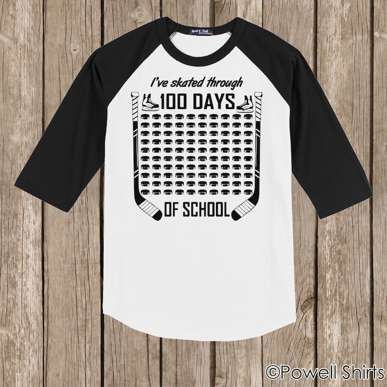 K&ouml;nnte beinhalten: Ein wei&szlig;-schwarzes Baseball-T-Shirt mit einem Hockey-Puck-Design. Der Text auf dem Shirt lautet "I've skated through 100 days of school".