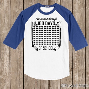 K&ouml;nnte beinhalten: Wei&szlig;es und blaues Baseball-T-Shirt mit einem Hockey-Puck-Design. Das Shirt sagt "Ich bin 100 Tage lang durch die Schule gelaufen".