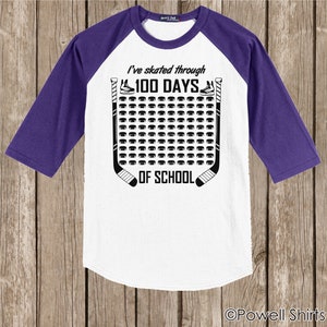 K&ouml;nnte beinhalten: Wei&szlig;es und violettes Baseball-T-Shirt mit einem Hockey-Puck-Design. Das Shirt sagt "Ich bin 100 Tage lang durch die Schule gelaufen".