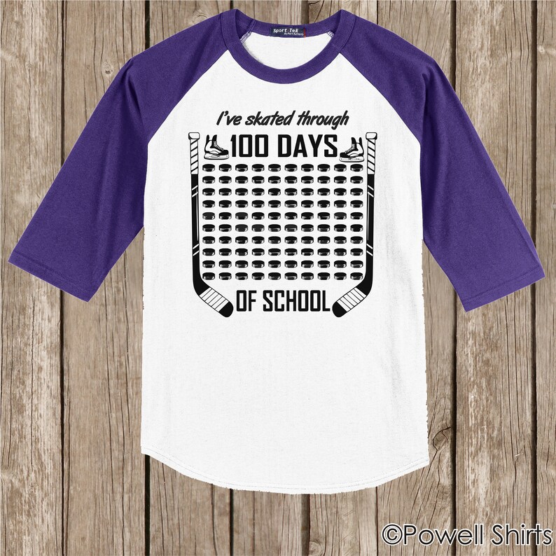 K&ouml;nnte beinhalten: Wei&szlig;es und violettes Baseball-T-Shirt mit einem Hockey-Puck-Design. Das Shirt sagt "Ich bin 100 Tage lang durch die Schule gelaufen".