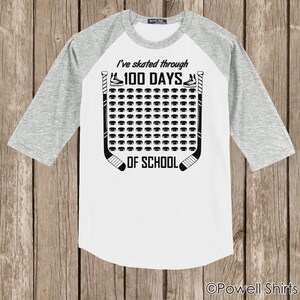 K&ouml;nnte beinhalten: Ein wei&szlig;es und graues Baseball-T-Shirt mit einem Hockey-Motiv. Das Shirt zeigt eine Grafik mit Hockeyschl&auml;gern und Pucks mit dem Text "Ich bin 100 Tage lang durch die Schule gelaufen".
