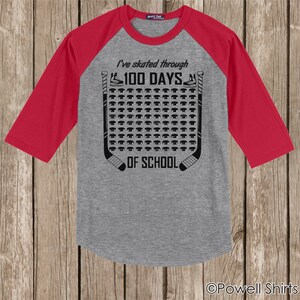 K&ouml;nnte beinhalten: Ein graues und rotes Baseball-T-Shirt mit einem Hockey-Puck-Design. Das Shirt sagt "Ich bin 100 Tage durch die Schule gelaufen".