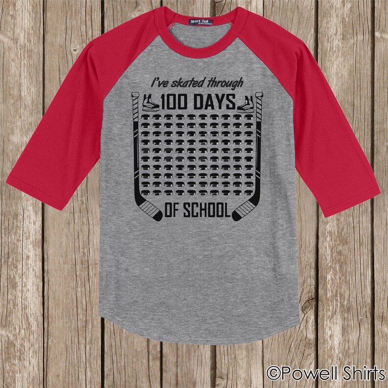 K&ouml;nnte beinhalten: Ein graues und rotes Baseball-T-Shirt mit einem Hockey-Puck-Design. Das Shirt sagt "Ich bin 100 Tage durch die Schule gelaufen".