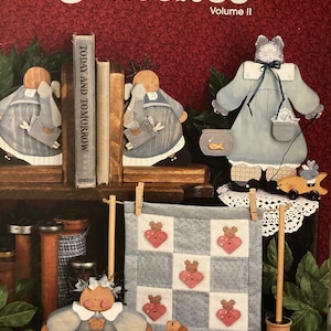 May include: A book titled "Patches Volume II" with a red background. The book features illustrations of painted wooden figures, including angels, a cat, and a girl holding a teddy bear. The book also includes a quilt pattern with hearts.