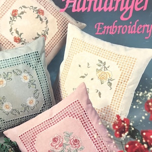 May include: Four embroidered cushions with floral patterns in pink, cream, and blue-green. Each cushion features detailed openwork borders. The image includes the text "An Introduction to Hardanger Embroidery".