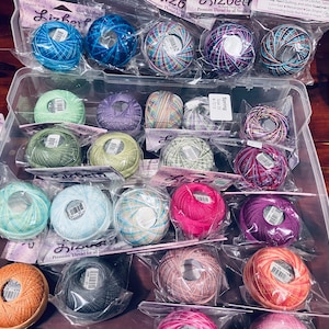 May include: A close-up of a variety of colourful embroidery floss spools in plastic packaging. The spools are arranged in rows and labelled with the brand name "Lizbeth" and the size "20".