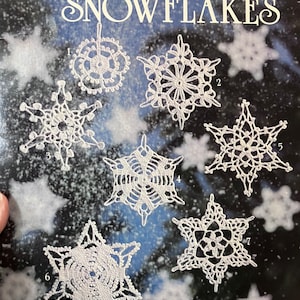 May include: Crocheted snowflake patterns from the American School of Needlework book "Crocheted Snowflakes" book number 1025(25) by Mary Thomas. The book cover features seven different snowflake designs.