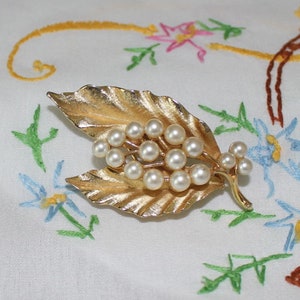 May include: A gold-toned leaf brooch with a cluster of faux pearls. The brooch is on a white fabric background with embroidered flowers.