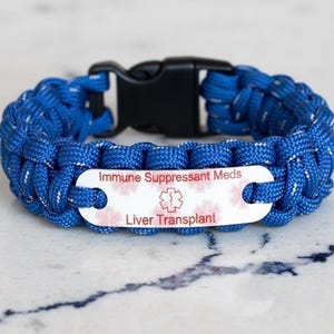 May include: A blue paracord medical alert bracelet featuring a black plastic side-release buckle and a white metal identification plate. The plate displays the text Immune Suppressant Meds and Liver Transplant with a red Star of Life symbol.