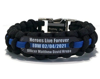 May include: A black paracord bracelet featuring a central metal plate with a thin blue line. The plate is engraved with the text: Heroes Live Forever, EOW 02/04/2021, Officer Matthew David Krupa. It includes a silver-toned metal buckle.