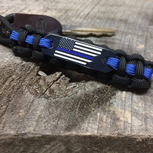 May include: Black and blue paracord keychain with a metal tag featuring the American flag and a blue line. The keychain is attached to a key and sits on a weathered wooden surface.