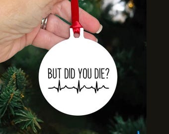 May include: White, round Christmas ornament with the text "BUT DID YOU DIE?" in black, along with a black heartbeat line. The ornament is held by a hand with a red ribbon for hanging. The background is a blurred Christmas tree.