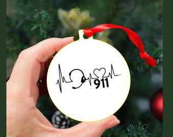 May include: White round Christmas ornament with a red ribbon. The ornament features a black graphic of a heartbeat, a headset, a heart, and the number 911. The ornament is held in a hand.