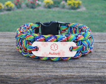 May include: A medical alert bracelet made of rainbow-colored paracord with a black plastic side-release buckle. The bracelet features a white metal identification plate with a red Star of Life symbol and the word Autism printed in red text.