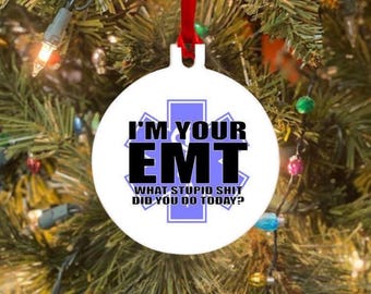 May include: White round Christmas ornament with the text "I'M YOUR EMT WHAT STUPID SHIT DID YOU DO TODAY?" in black and white. The ornament has a red ribbon for hanging and is set against a backdrop of a Christmas tree.