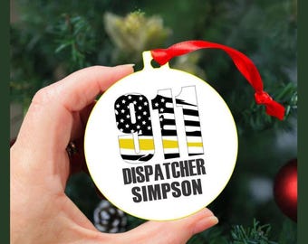 May include: A white round Christmas ornament with the text "911 DISPATCHER SIMPSON". The numbers 911 are in black and white with a yellow stripe. A red ribbon is attached for hanging.