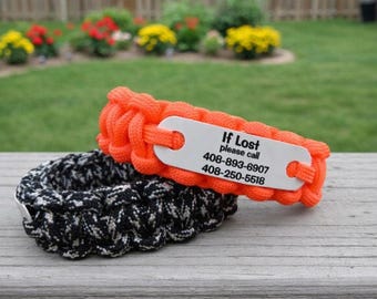 May include: Two paracord collars featuring a braided design. The top collar is bright orange with a silver-toned metal identification tag engraved with the text: If Lost please call 408-893-6907 408-250-5518. The bottom collar is black and grey.