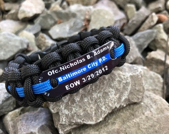 Puede incluir: Pulsera de paracord negro con una raya azul y una etiqueta de metal que dice "Ofc. Nicholas B. Adams Baltimore City P.D. EOW 3/29/2012".