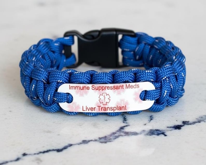 May include: A blue paracord medical alert bracelet featuring a black plastic side-release buckle and a white metal identification plate. The plate displays the text Immune Suppressant Meds and Liver Transplant with a red Star of Life symbol.