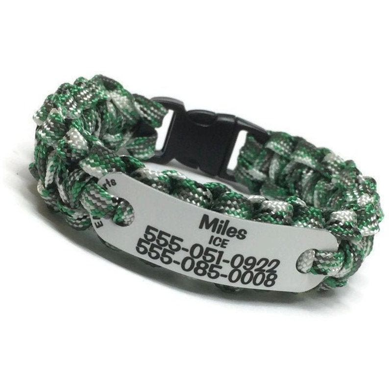 May include: A paracord bracelet featuring a braided pattern in green, grey, and black. It includes a black plastic side-release buckle and a silver-toned metal identification plate engraved with the name Miles, ICE, and two phone numbers: 555-051-0922 and 555-085-0008.